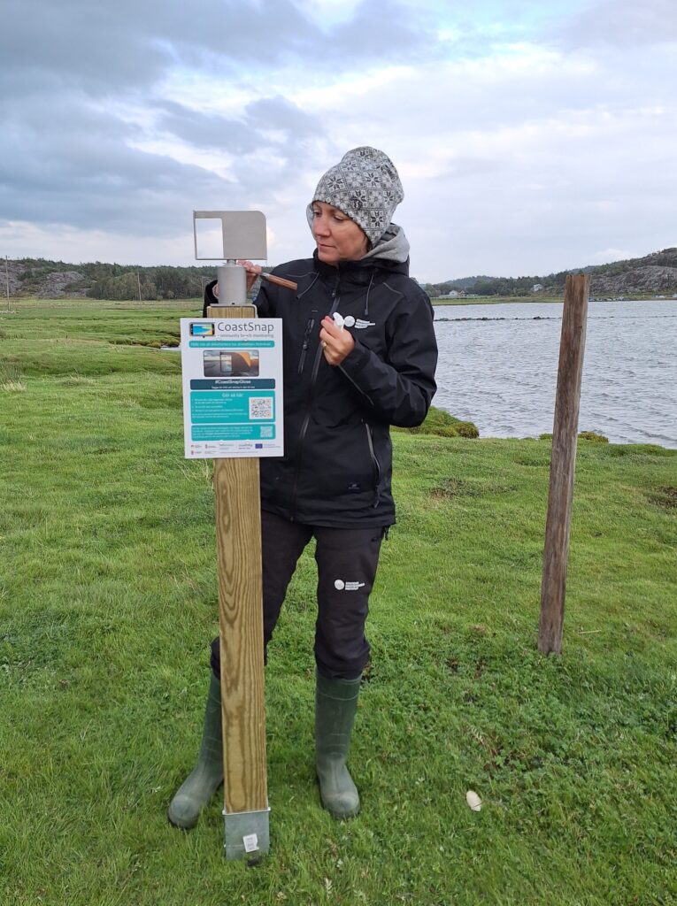 En kvinna med svarta friluftskläder och gröna stövlar står på en strandäng och skrapar med en morakniv på ett fotostativ. 