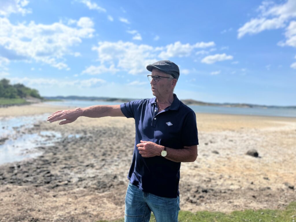 Man i blå t-shirt och keps står på en strand och pekar med högerhanden mot ett osynligt mål. 